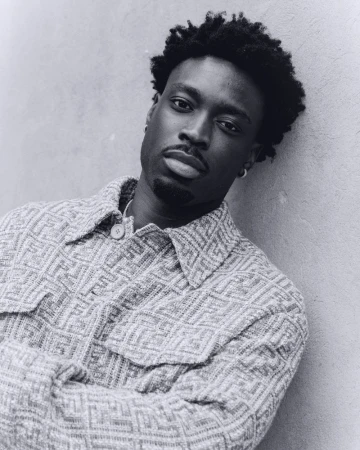 Portrait of a Black man with short curly hair, wearing a patterned button-down shirt, leaning against a gray wall indoors.