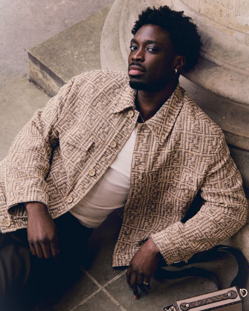 Stylish African-American man poses on a concrete ledge wearing a patterned jacket, white t-shirt, and dark pants, with a shoulder bag.