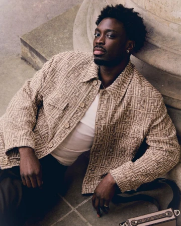 Stylish African-American man poses on a concrete ledge wearing a patterned jacket, white t-shirt, and dark pants, with a shoulder bag.