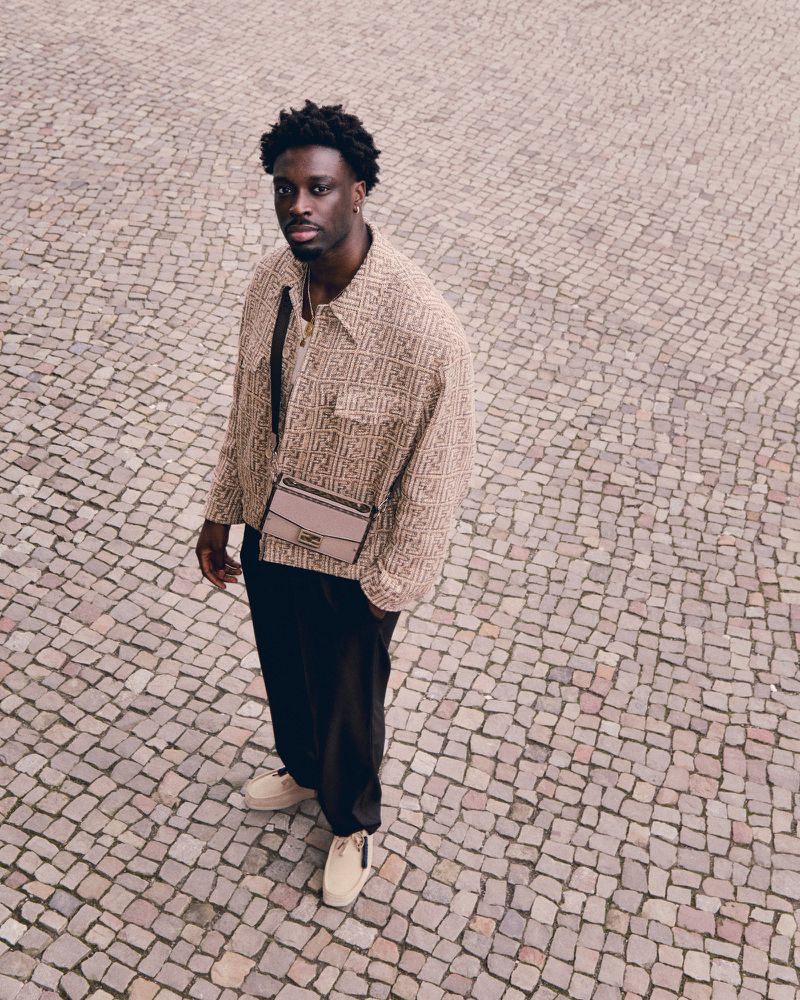 A stylish Black man stands on a stone-paved surface wearing a patterned jacket, black pants, beige shoes, and a cross-body bag.
