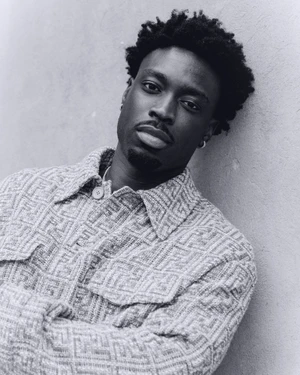 Portrait of a Black man with short dark hair wearing a patterned button-down jacket, leaning against a textured wall.