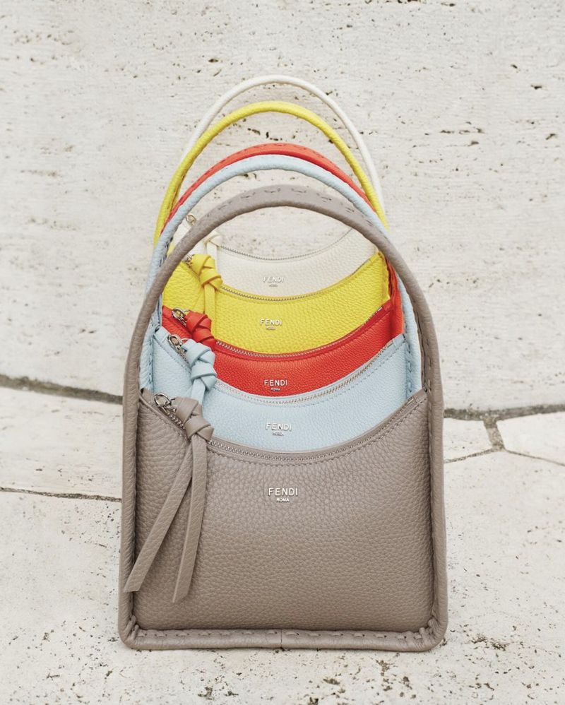 Stack of colorful Fendi handbags in yellow, orange, blue, and taupe leather, arranged vertically on a travertine surface.