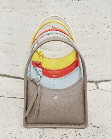 Stack of colorful Fendi handbags in yellow, orange, blue, and taupe leather, arranged vertically on a travertine surface.