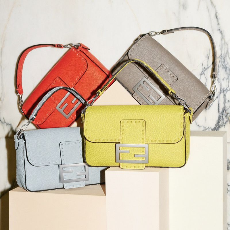 Four Fendi handbags in different colors: red, gray, yellow, and light blue. The handbags are arranged on a two-tiered platform.