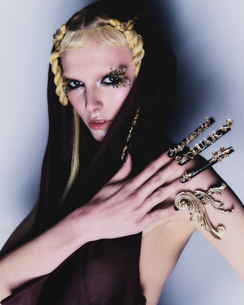 Eye-level portrait of a woman with blonde braided hair and dramatic eye makeup wearing a hooded brown top in a light studio.