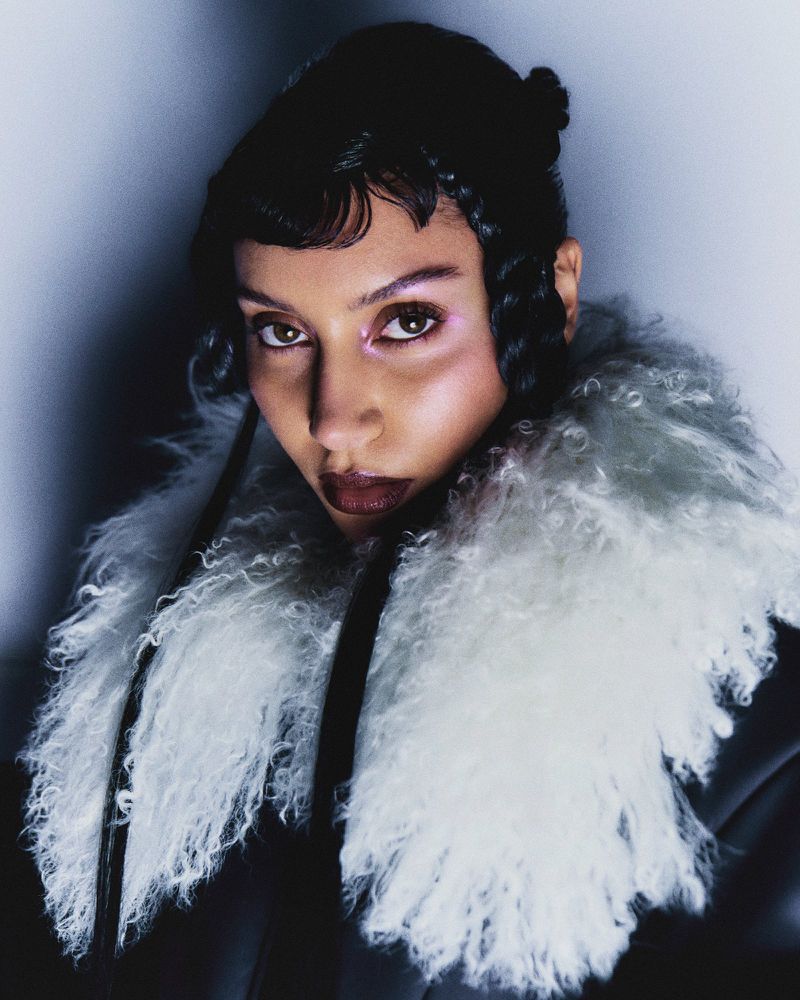 Close-up portrait of a woman with dark hair and fair skin, wearing a textured faux fur collar, against a soft, diffused light.