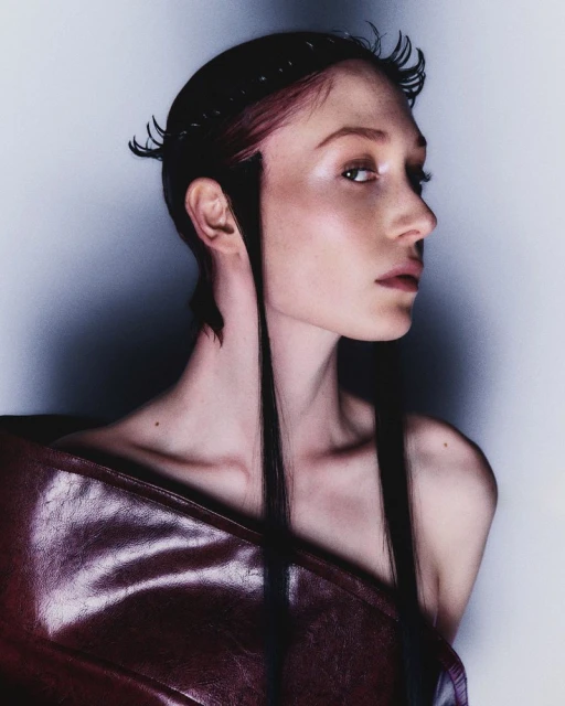 Portrait of a woman with long, straight dark hair and an unconventional hairstyle wearing a burgundy, glossy, draped garment.