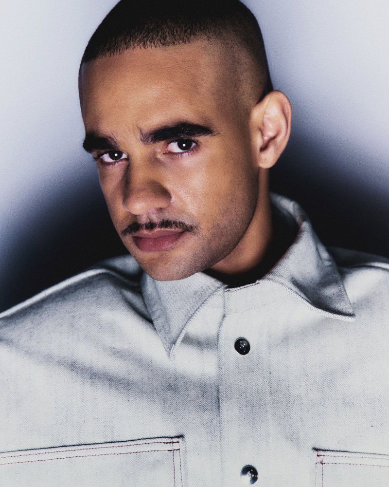 Portrait of an adult male with a mustache, wearing a light-colored denim button-down shirt against a gradient grey background.