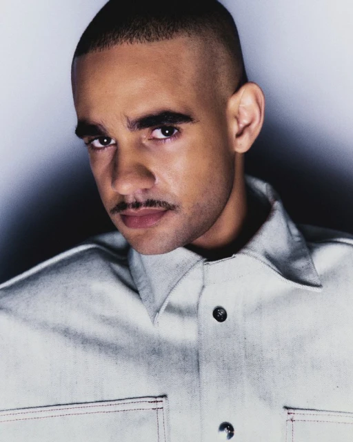 Portrait of an adult male with a mustache, wearing a light-colored denim button-down shirt against a gradient grey background.