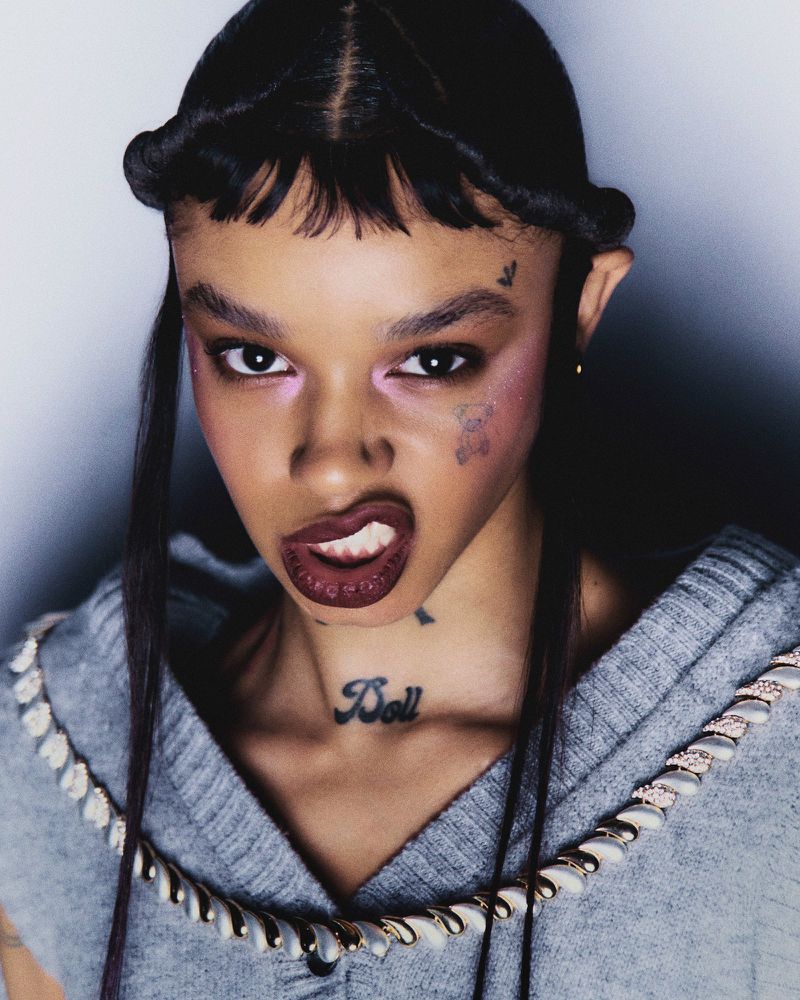 Close-up portrait of a young adult female model with unique braided hairstyle, distinctive face tattoos, wearing a grey top, and dark lipstick.