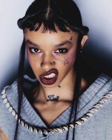 Close-up portrait of a young adult female model with unique braided hairstyle, distinctive face tattoos, wearing a grey top, and dark lipstick.