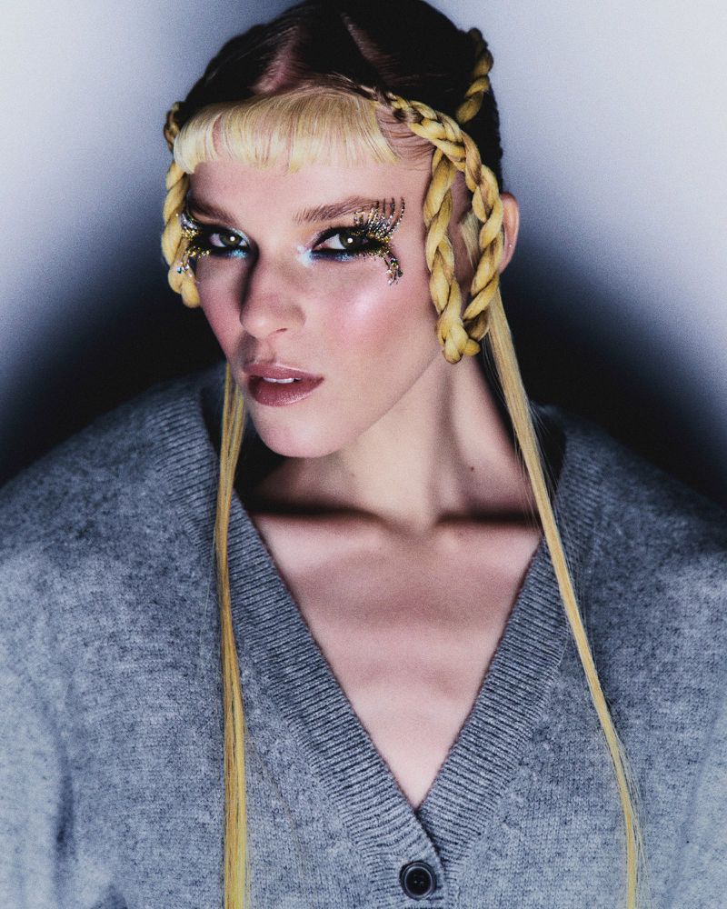 Close-up shot of a woman with blonde hair, creative eye makeup, and a gray cardigan against a gradient white to dark gray background.