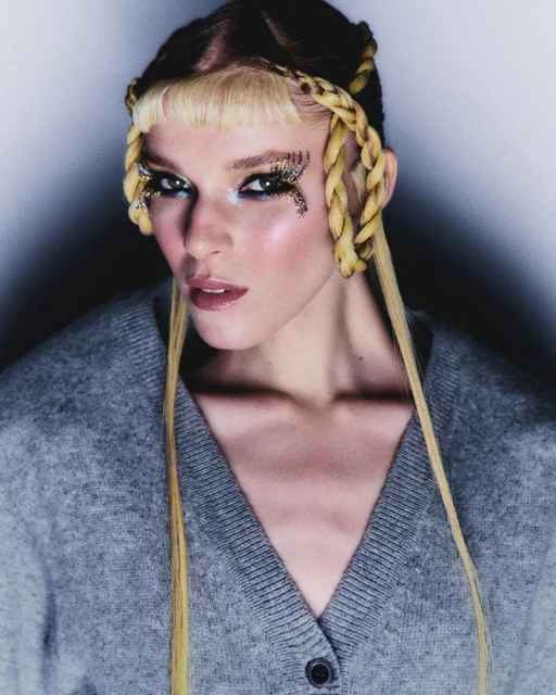 Close-up shot of a woman with blonde hair, creative eye makeup, and a gray cardigan against a gradient white to dark gray background.