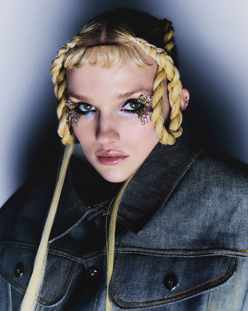 Eye-level portrait of a young adult female wearing a denim jacket with a braided blond hairstyle and glitter eye makeup in a grey-toned studio.