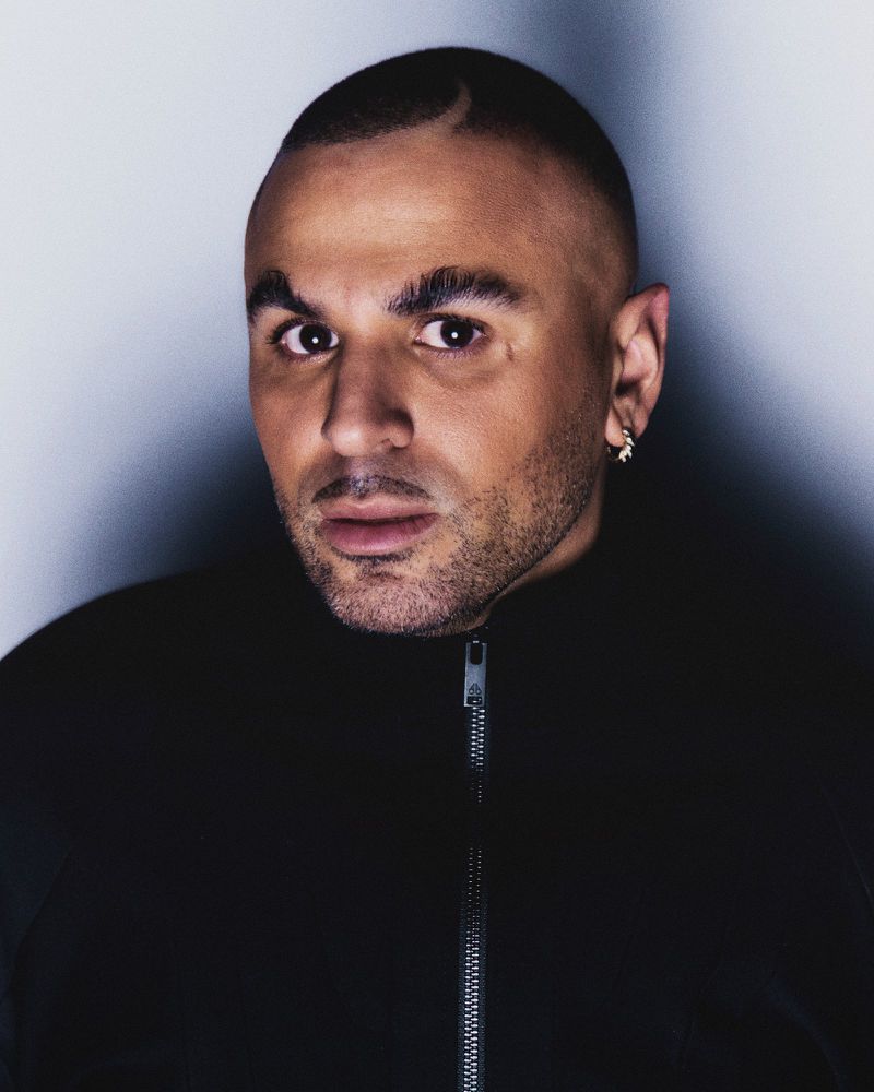 Close-up portrait of a man with a shaved head and an earring, wearing a black jacket against a neutral gray backdrop.