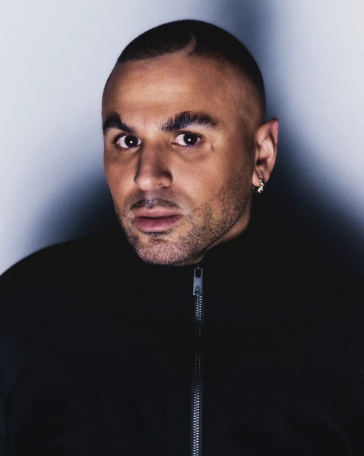 Close-up portrait of a man with a shaved head and an earring, wearing a black jacket against a neutral gray backdrop.