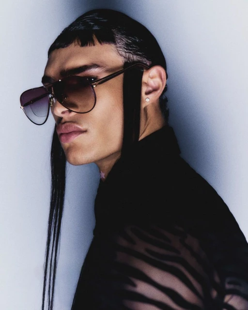A close-up studio shot features a person with slicked-back hair, wearing sunglasses and a sheer patterned top, against a neutral backdrop.