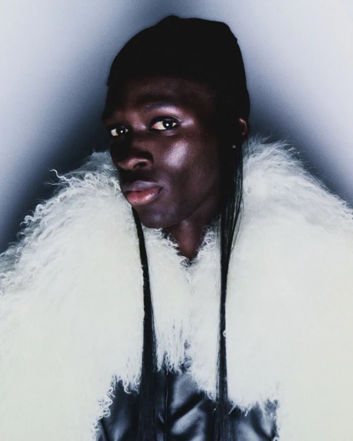 Close-up portrait of a dark-skinned person with long hair wearing a white, furry coat against a gradient background.