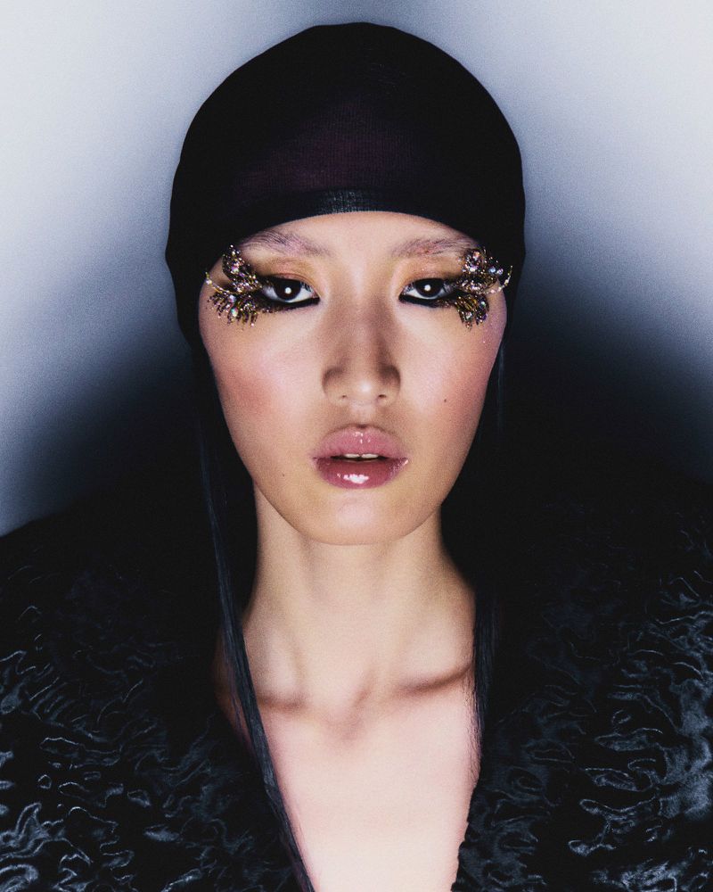 Close-up portrait of an Asian woman with dramatic eye makeup, wearing a black headscarf and textured top against a dark background.