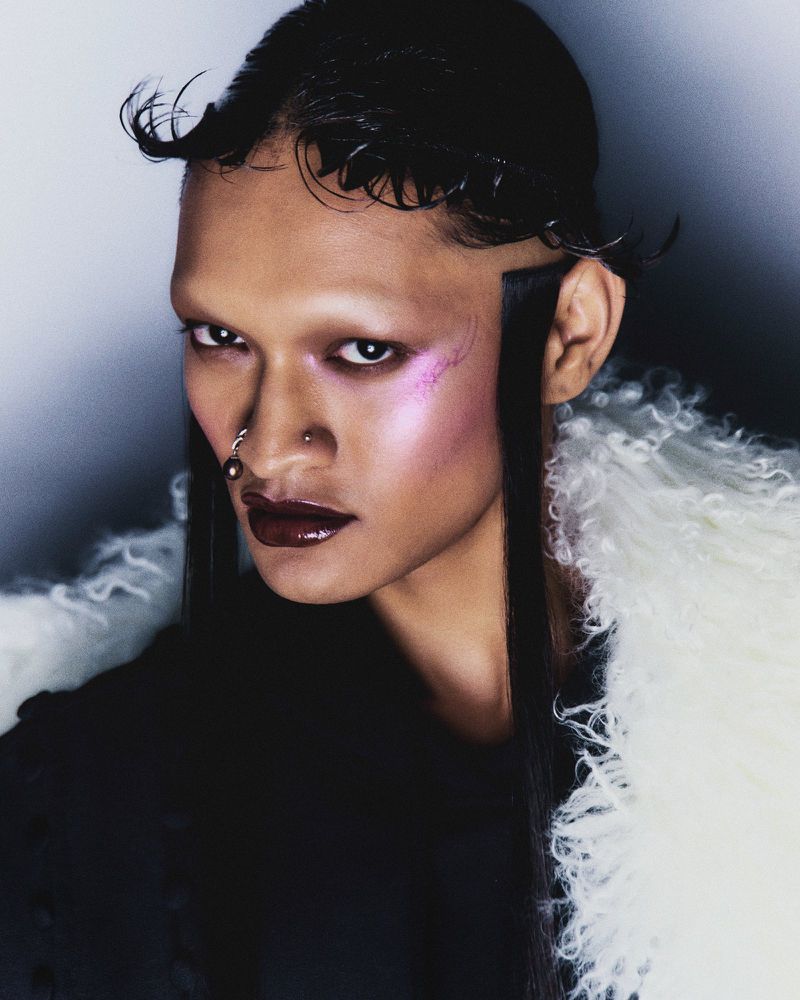 Androgynous person with dark hair and dark lipstick wearing white fur collar, posing in a studio with bright lighting and a dark background.