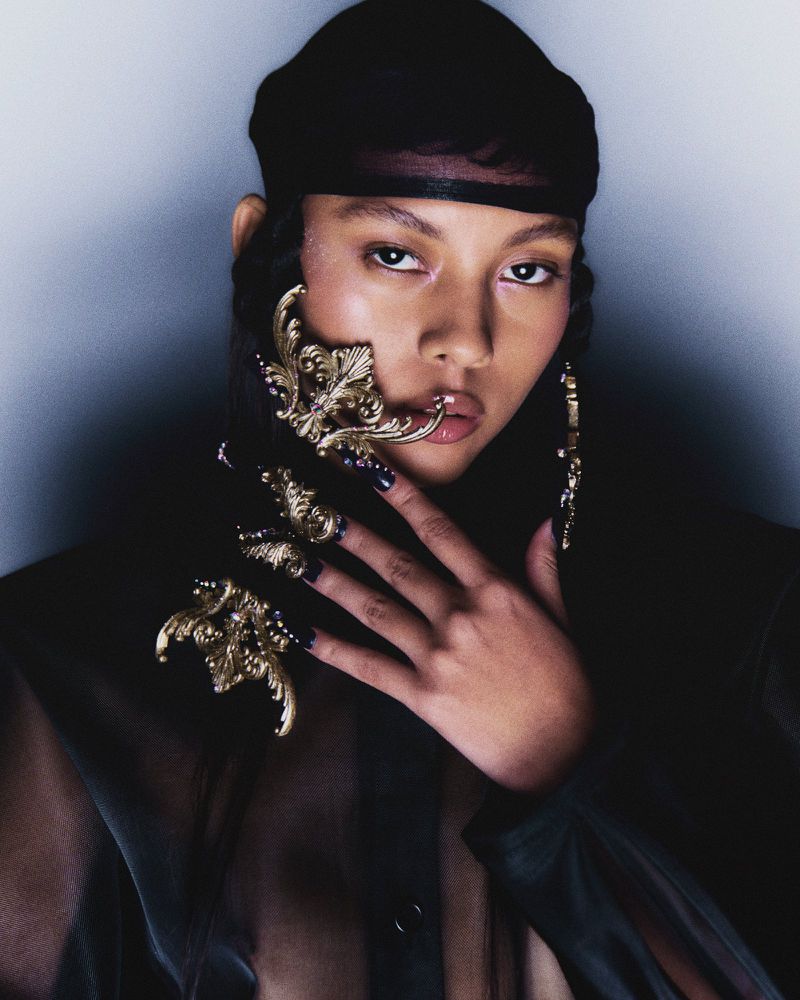 Headshot of a young adult woman in a sheer black top and headscarf, posing with ornate gold hand and face jewelry against a neutral wall.