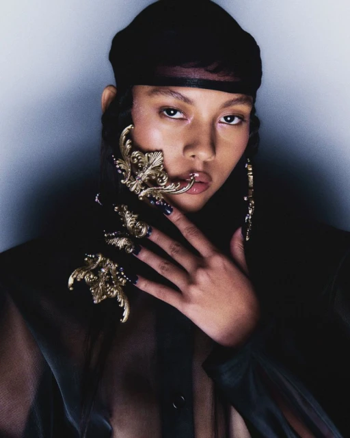 Headshot of a young adult woman in a sheer black top and headscarf, posing with ornate gold hand and face jewelry against a neutral wall.