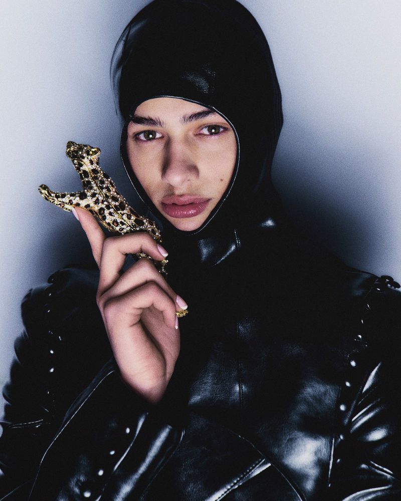 Close-up of a person in a black hooded outfit holding a gold leopard figurine against a gray backdrop, studio lighting.