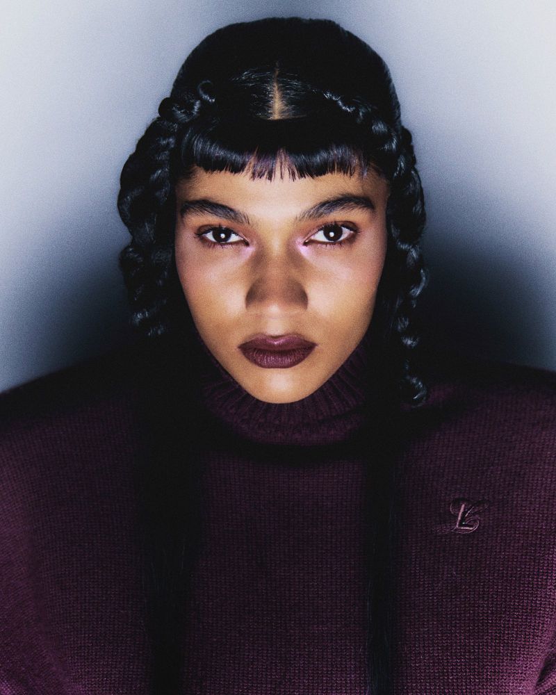 Close-up portrait of an adult female with dark hair styled with braids, wearing a burgundy turtleneck sweater in a studio setting.
