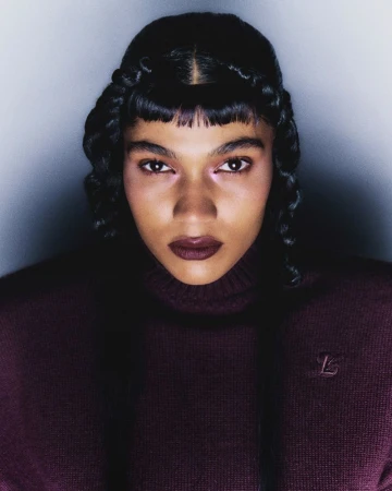 Close-up portrait of an adult female with dark hair styled with braids, wearing a burgundy turtleneck sweater in a studio setting.