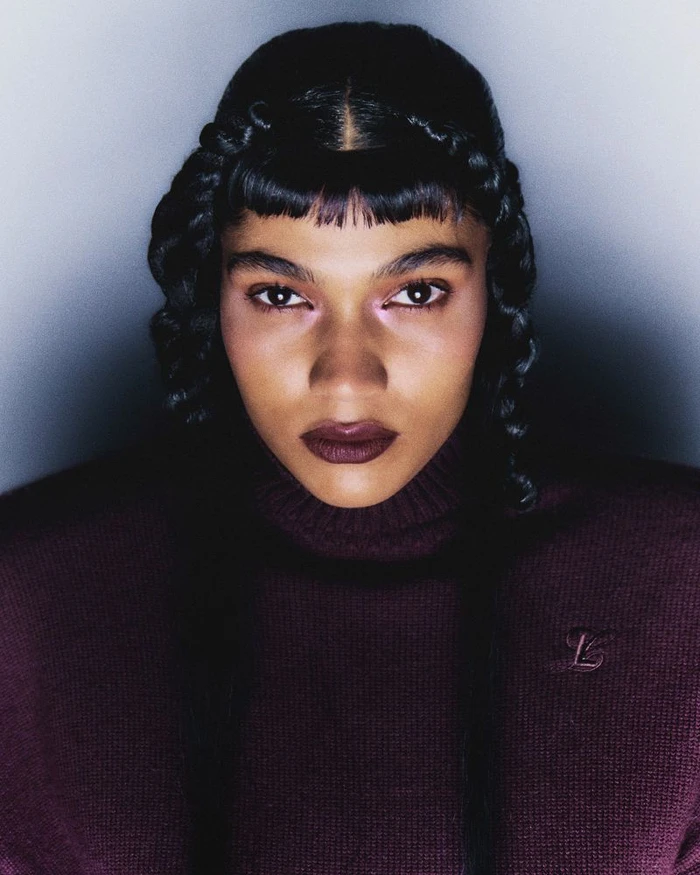 Close-up portrait of a model with intricate braided hair and dark plum lipstick wearing a plum turtleneck sweater against a vignette background.