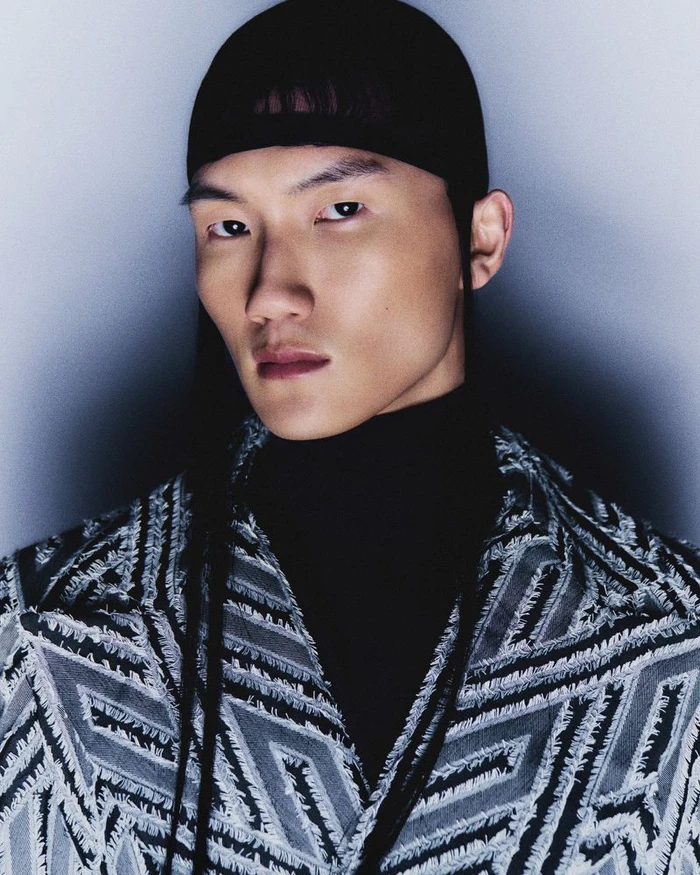 Portrait of an East Asian male model wearing a patterned textured jacket and black cap against a light background