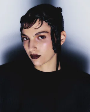 Close-up portrait of a model with dark braided hair, dark lipstick, and dark top against a soft gradient background.