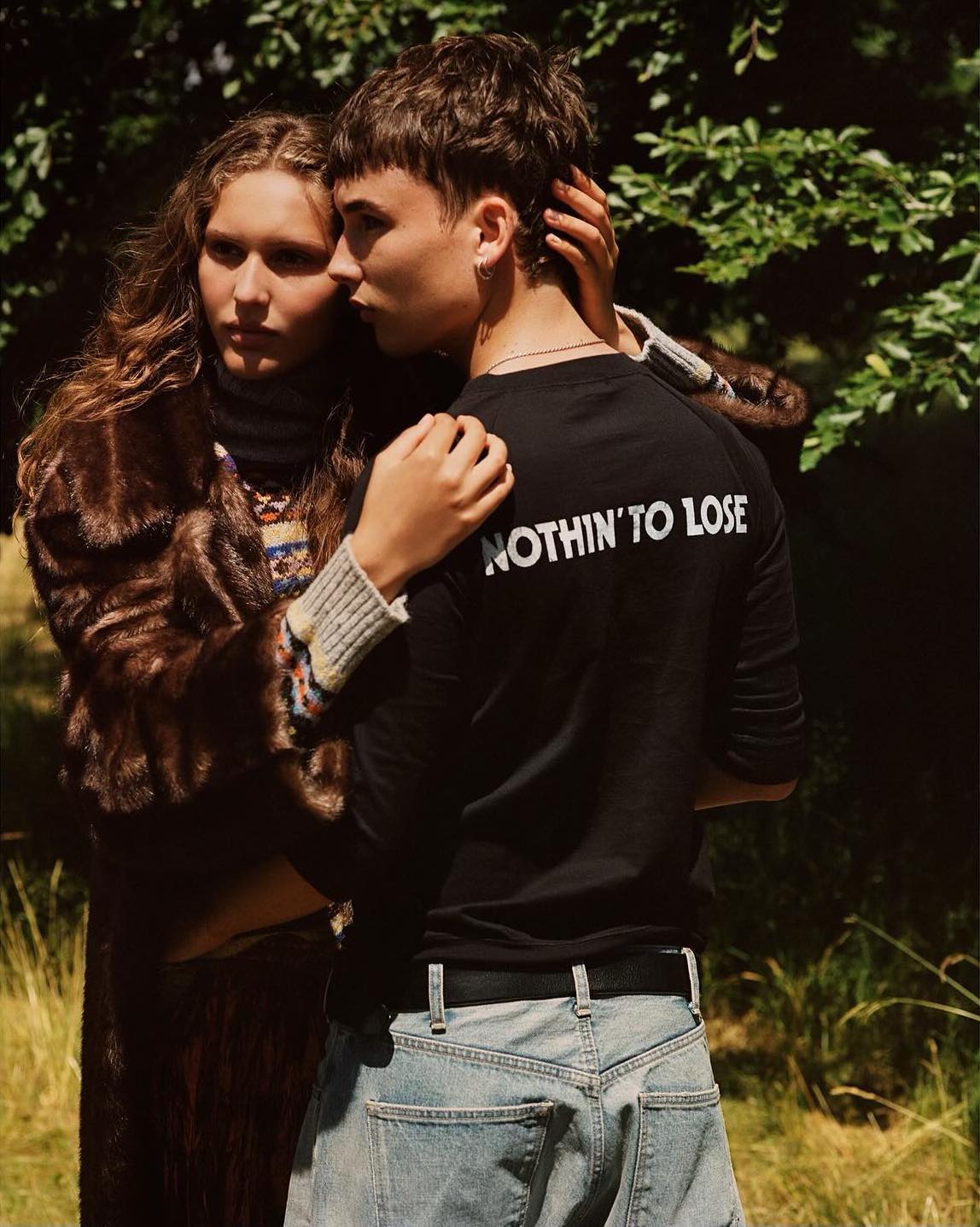 A woman in a fur coat poses with a man in a black t-shirt with the text, NOTHIN' TO LOSE, in an outdoor setting with green foliage in the background.