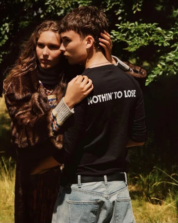 A woman in a fur coat poses with a man in a black t-shirt with the text, NOTHIN' TO LOSE, in an outdoor setting with green foliage in the background.