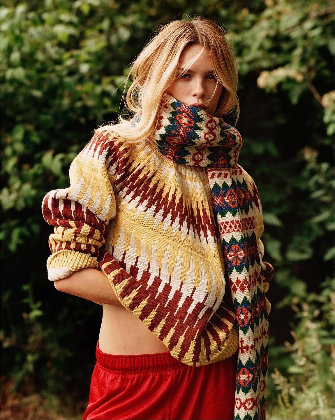 Adult woman with fair hair poses in a patterned sweater and scarf, wearing red velvet shorts outdoors against a leafy green background.