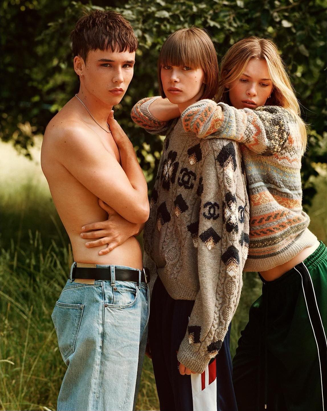 Three young adults pose outdoors. One shirtless man stands beside two women wearing knit sweaters, with lush green foliage behind them.
