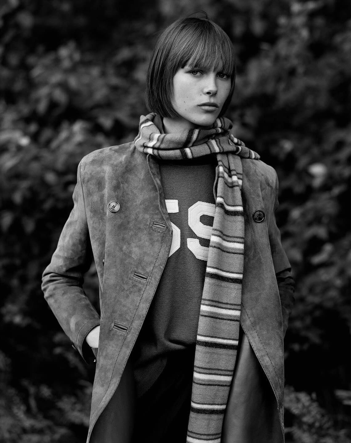 Black and white portrait of a young woman with bangs wearing a suede coat, sweater, and striped scarf, posing outdoors against a leafy background.