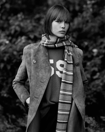 Black and white portrait of a young woman with bangs wearing a suede coat, sweater, and striped scarf, posing outdoors against a leafy background.