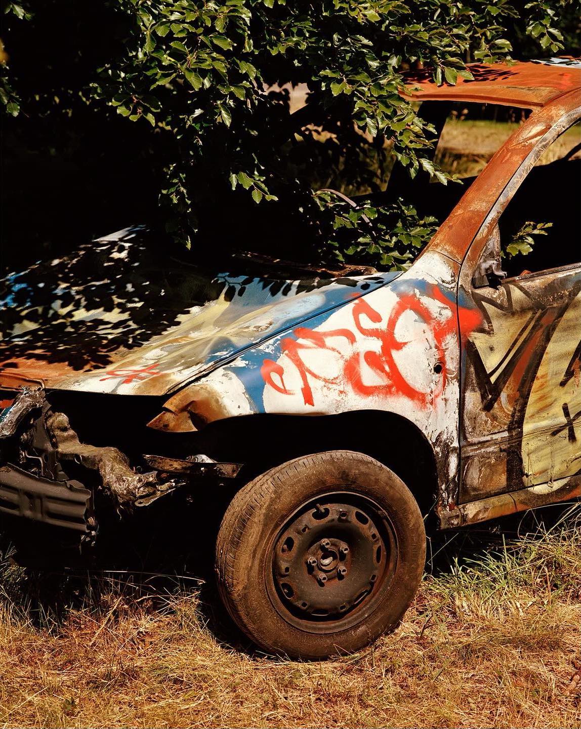 Wrecked car with graffiti in an outdoor setting, surrounded by trees and grass, showing damage and rust on the vehicle.