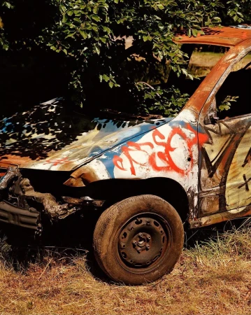 Wrecked car with graffiti in an outdoor setting, surrounded by trees and grass, showing damage and rust on the vehicle.
