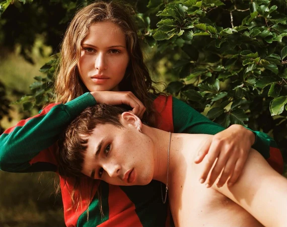 Young woman resting her hands and chin on the shoulder of a shirtless young man amidst lush green foliage.