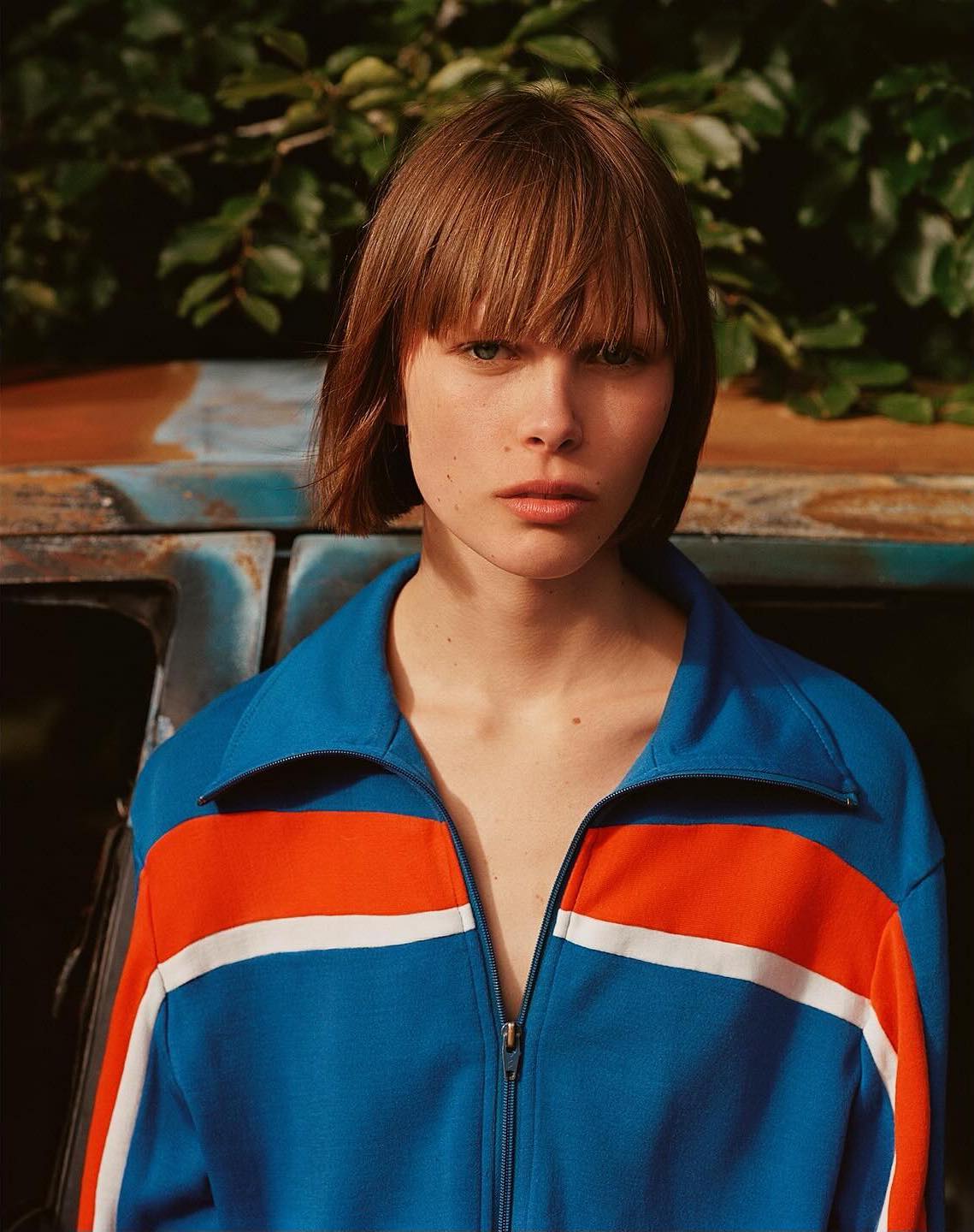 Portrait of a woman with a bob haircut wearing a blue and orange tracksuit jacket. Outdoor shot with natural light and green foliage.