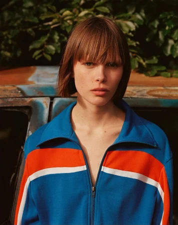 Portrait of a woman with a bob haircut wearing a blue and orange tracksuit jacket. Outdoor shot with natural light and green foliage.