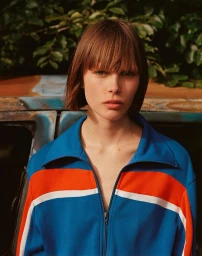 Model with bob haircut in a blue, red, and white track jacket against an outdoor rustic vehicle and green foliage background.