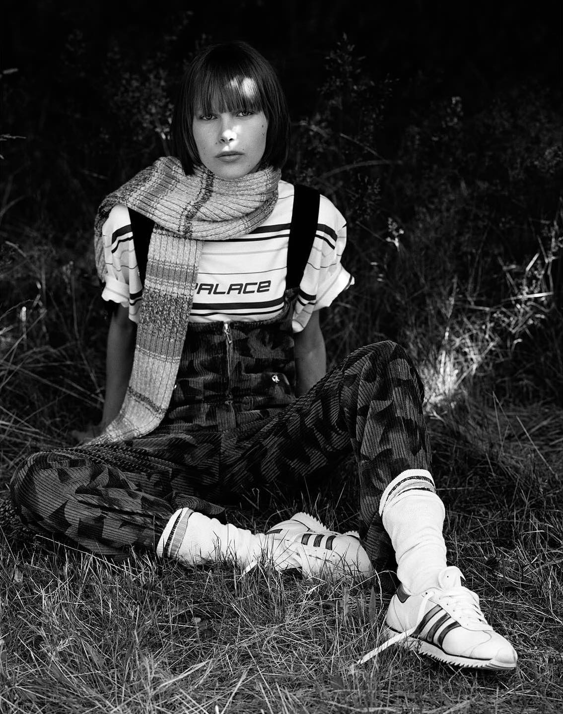 A young woman with bangs, wearing a scarf, t-shirt, and patterned pants, poses sitting in a grassy outdoor environment in black and white.