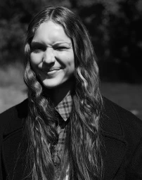 Monochrome portrait of a smiling woman with long wavy hair wearing a dark coat and collared shirt.