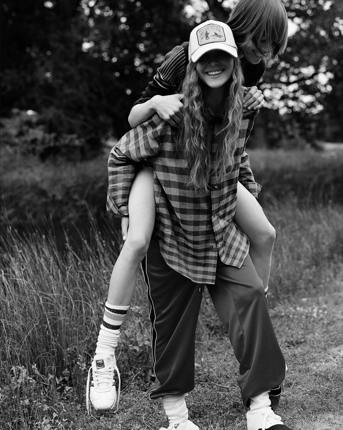 Two women in outdoor setting; one gives the other a piggyback ride with trees in the background, black and white photography.