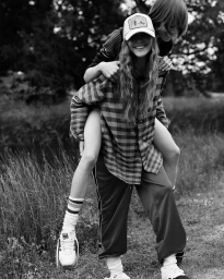 Black and white editorial photo of a young woman giving another person a piggyback ride outdoors in a park.