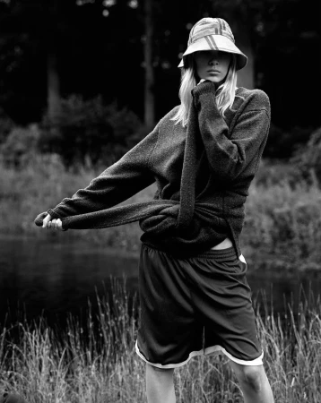 Black and white portrait of a model wearing a bucket hat and sweater standing outdoors by tall grass