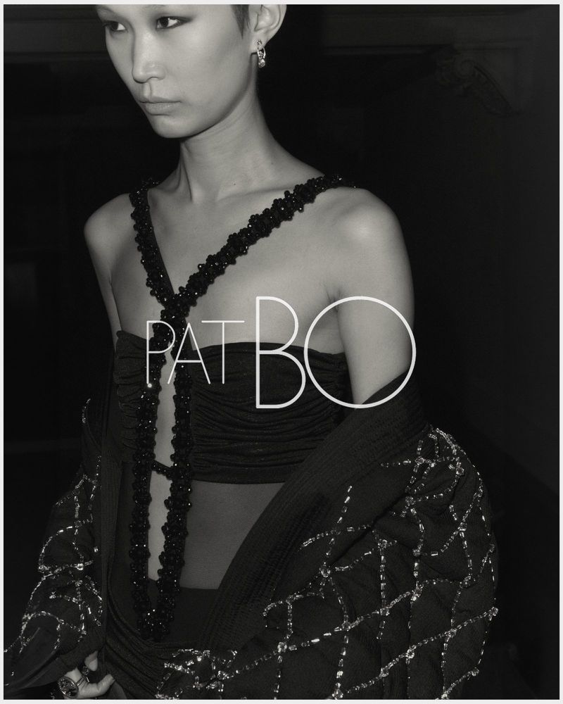 Black and white fashion shot of an Asian female model wearing a jeweled top with the PatBO logo. Shot in a dark environment.